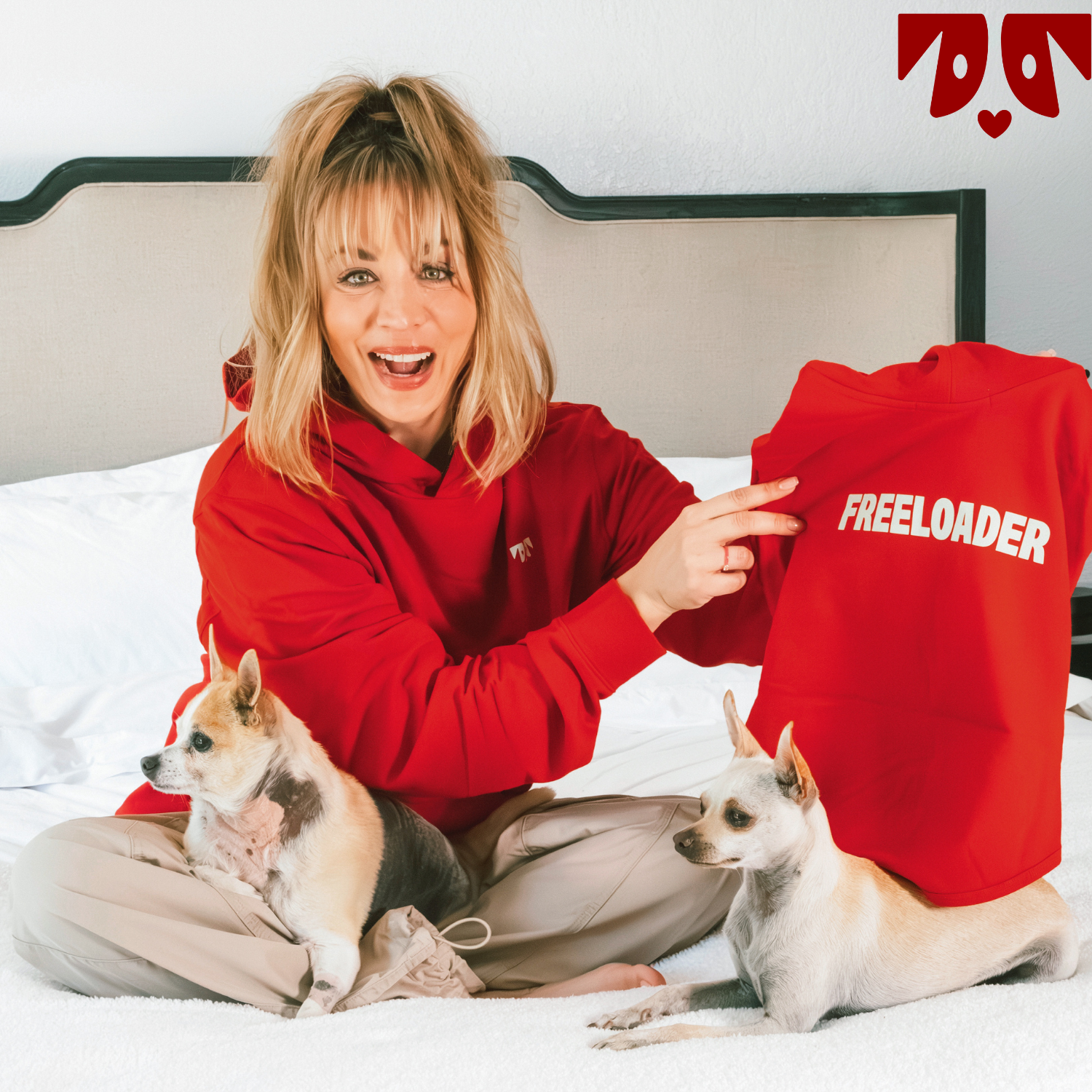 Kaley Cuoco in red hoodie sitting on floor with two dogs, holding up  a red shirt labeled 'FREELOADER'
