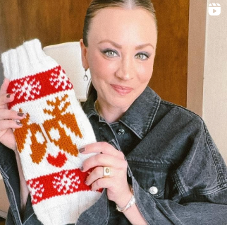 Kaley Cuoco holding a white and red knitted item with a reindeer pattern, smiling at the camera.