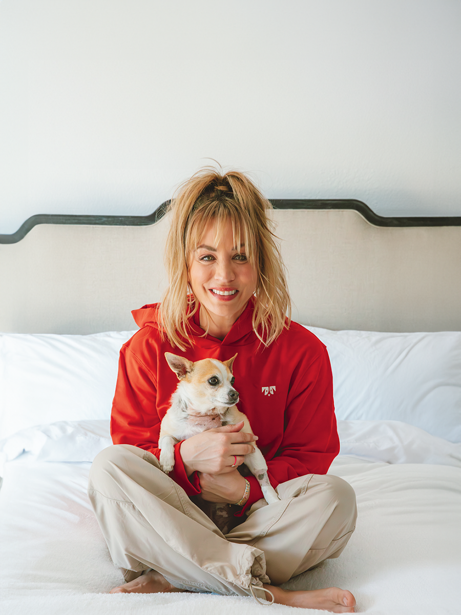 Kaley Cuoco in red hoodie sitting cross-legged on bed, smiling and holding a small tan-and-white chihuahua