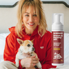 Woman in a red hoodie holding a small dog next to a bottle labeled 'OH NORMAN! Fresh & Foamy AF! Waterless Cleanser for Dogs No bath, no problem! 99.05% Natural Magic Mandarin'.