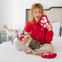 Actress Kaley Cuoco sitting on a bed holding a red heart-shaped recycled plush toy while cuddling one small dog and another small dog stands nearby