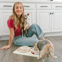 Cream ceramic pet bowls with red pattern on cream mat; small tan dog eating while actress Kaley Cuoco in red shirt holds a chihuahua.