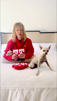 Actress Kaley Cuoco in red hoodie on a white bed with two small chihuahuas, showing a red heart-shaped recycled plush toy with white face design