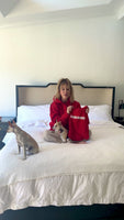 Front view of Caretaker Human Hoodie in red worn by actress Kaley Cuoco holding a small red dog hoodie labeled FREELOADER on a bed